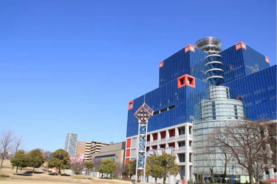 大阪儿童乐园KIDS PLAZA的独特现代建筑外观，蓝色与红色相间，在蔚蓝晴空下显得格外醒目，周围环绕着绿树，呈现宜人的景观。