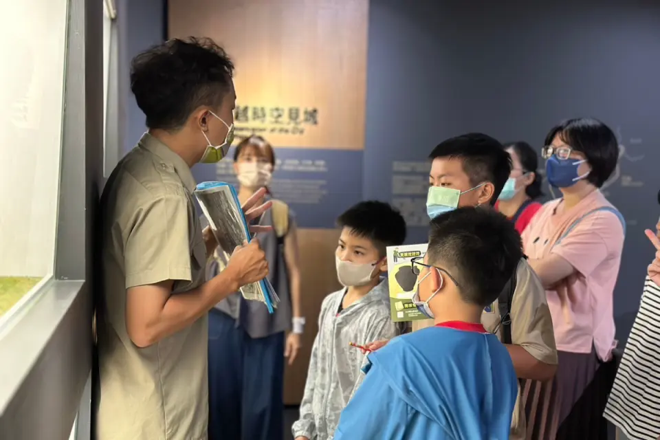A passionate guide explains exhibits inside a Zuoying cultural venue, with adults and children attentively listening, jointly exploring the wonderful stories of Kaohsiung Zuoying Old City and military villages.
