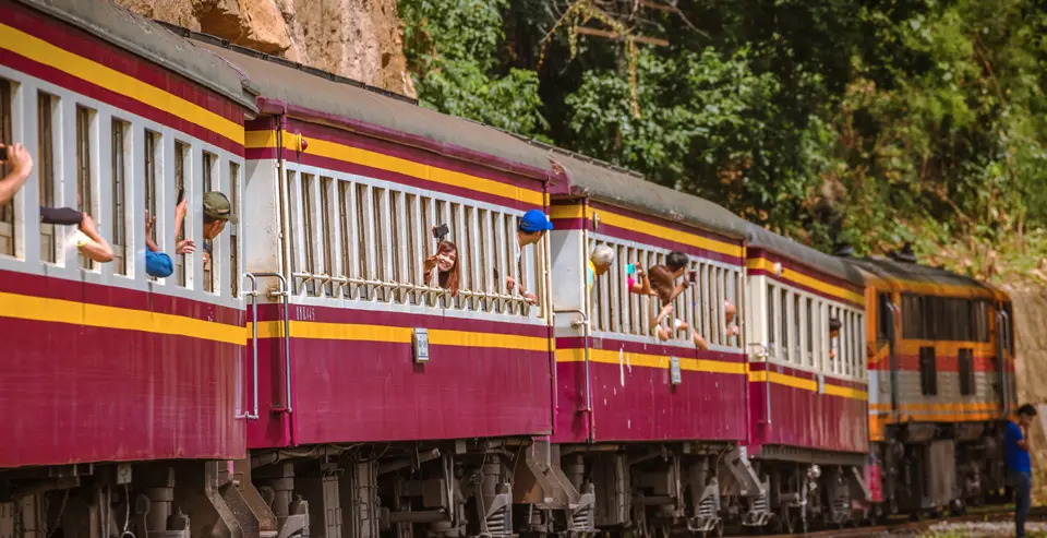 泰国北碧府包车一日游，体验乘坐火车沿着桂河畔的死亡铁路，乘客们探出头来欣赏沿途郁郁葱葱的山景。