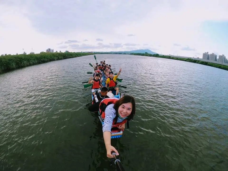 During a dragon boat experience on Taipei's Keelung River, a cheerful participant at the front captures a happy moment with teammates using a selfie stick, documenting their group water activity.