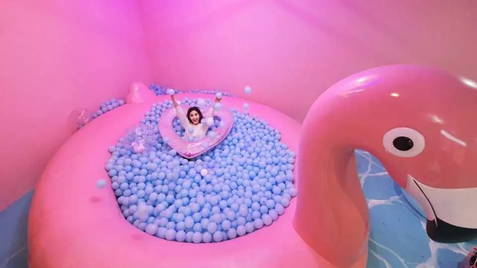 A woman enjoying a vibrant blue ball pit inside a giant pink flamingo float at Seoul's Insa-dong Color Pool Museum, capturing the fun, summer vacation atmosphere.