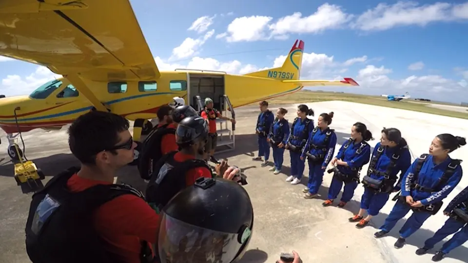 사이판 활주로에서 파란 점프슈트를 입은 참가자들이 노란 비행기 옆에서 강사로부터 안전 브리핑을 받으며 스릴 넘치는 스카이다이빙을 준비하는 모습.