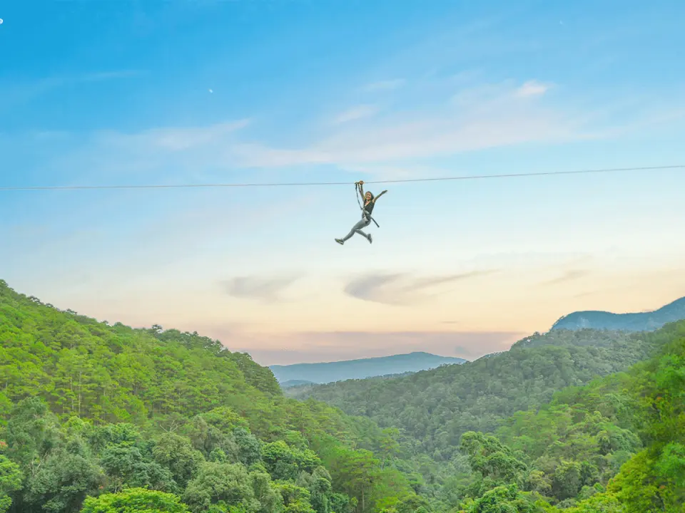 Một người đang trên zipline tại thác Datanla, Đà Lạt, Việt Nam, nhìn ra thung lũng xanh rộng lớn, trải nghiệm cảm giác bay lượn phấn khích trong ánh hoàng hôn.