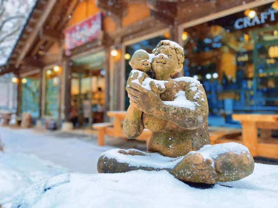 南怡島冬季雪中雕塑，溫馨的母子雕像被白雪覆蓋，背景是韓式建築與積雪的露天休息區。