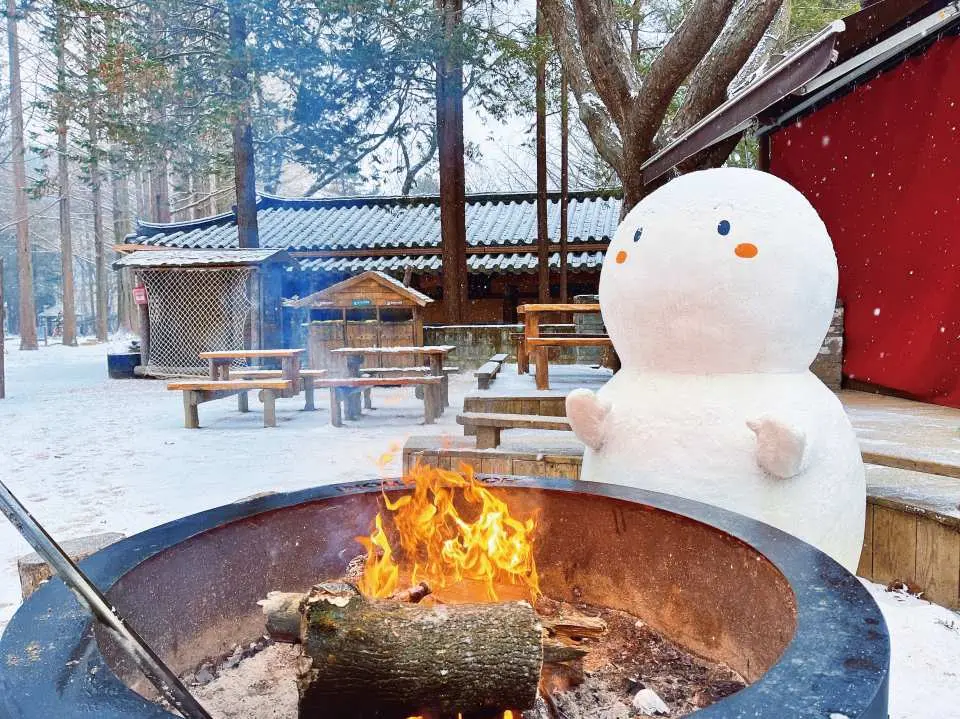 南怡島冬季暖意體驗，遊客在戶外火爐旁取暖，旁邊有可愛雪人相伴，感受溫馨的冬日氛圍。