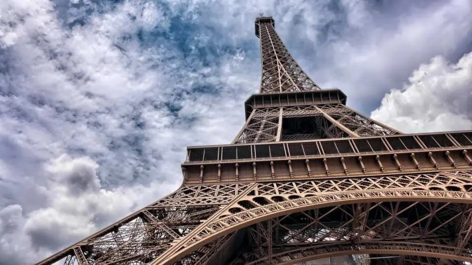 A dramatic low-angle view of the Eiffel Tower in Paris against a cloudy sky, showcasing its impressive iron structure and historical presence.