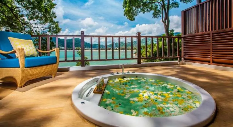Outdoor petal bathtub at Amari Phuket's Breeze Spa, a therapeutic spa bath overflowing with flowers, complemented by a wicker chair, offering serene sea views and blue skies for ultimate tranquility.