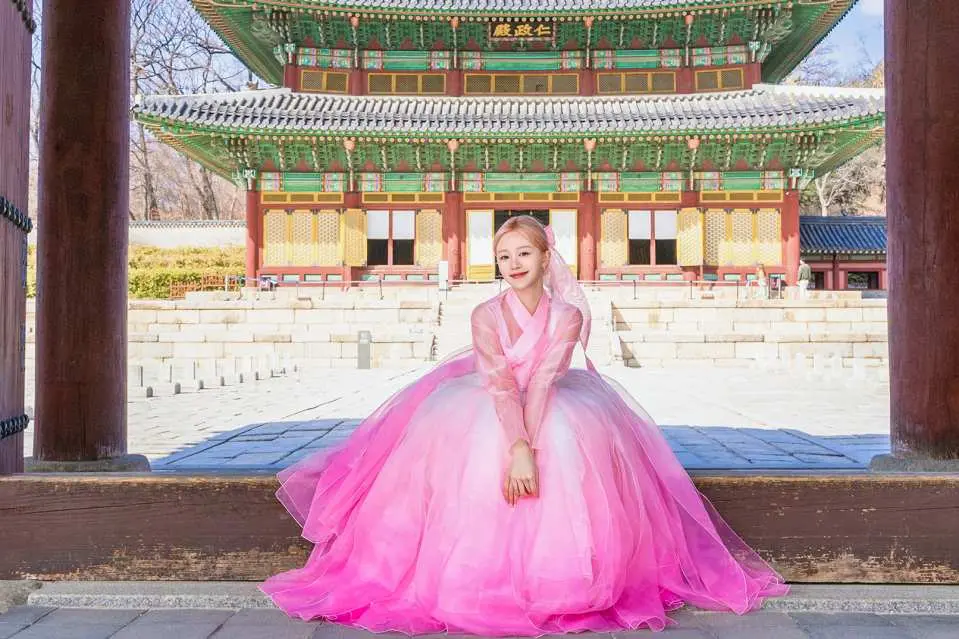 A woman in a beautiful pink gradient Hanbok elegantly seated in front of Seoul's majestic Changdeokgung Palace, enjoying a traditional Hanbok photoshoot experience.