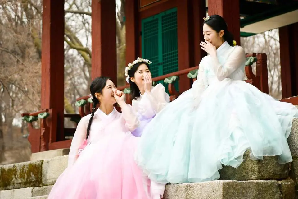 Three friends in colorful Hanboks laughing joyfully on the stone steps of a traditional palace in Seoul, sharing a memorable Hanbok photoshoot experience.