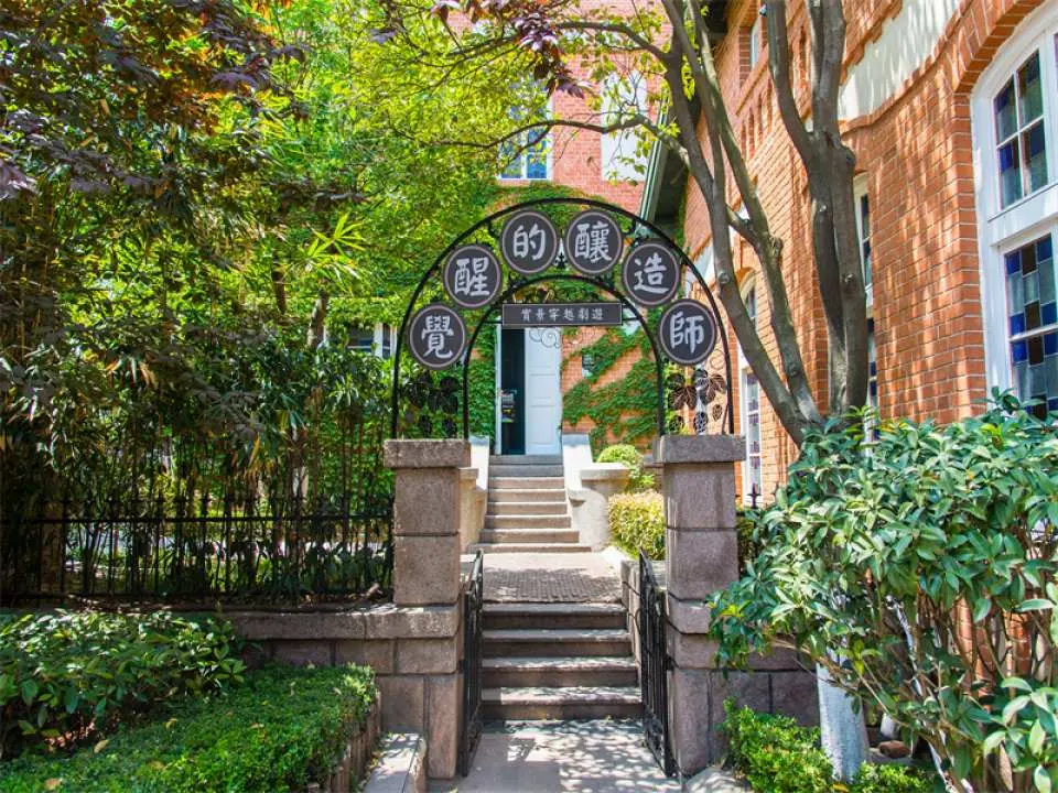 Lush garden entrance of Qingdao Beer Museum, featuring an ornate cast-iron archway inscribed with 'The Brewer,' inviting visitors to explore a century of beer legends.
