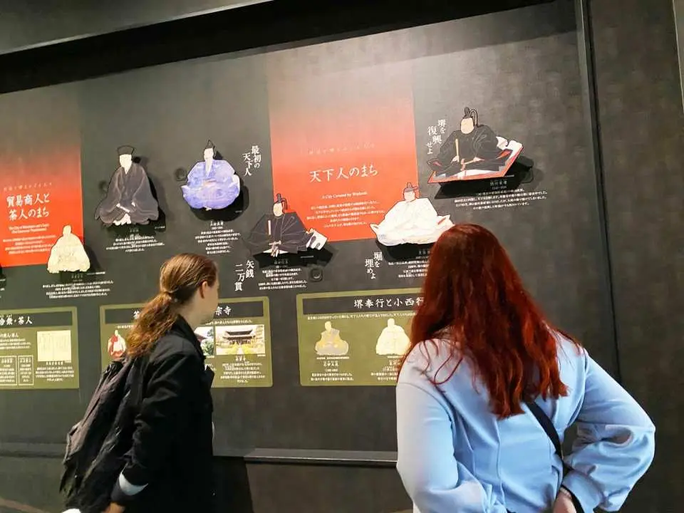 사카이 리쇼노모리 박물관에서 아즈치모모야마 시대 역사 인물과 다도 계승에 대한 전시 패널을 주의 깊게 관람하는 방문객들. 사카이 시의 풍부한 문화를 깊이 이해합니다.
