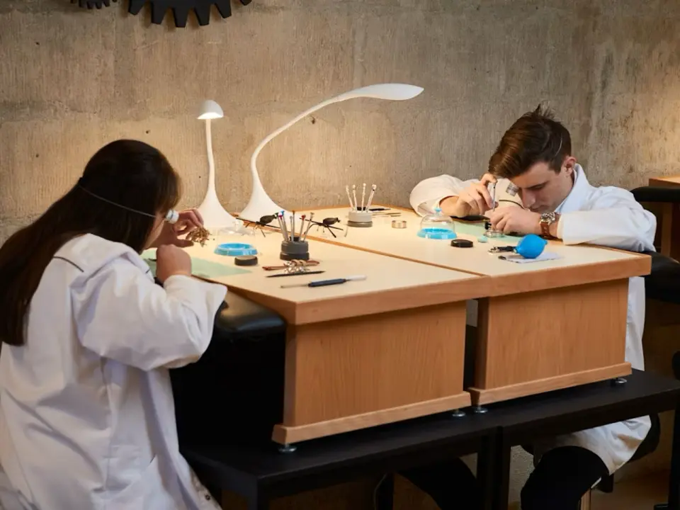 Two participants intently focused on assembling their watches at individual workbenches with specialized tools and lighting during an immersive Geneva watchmaking workshop.