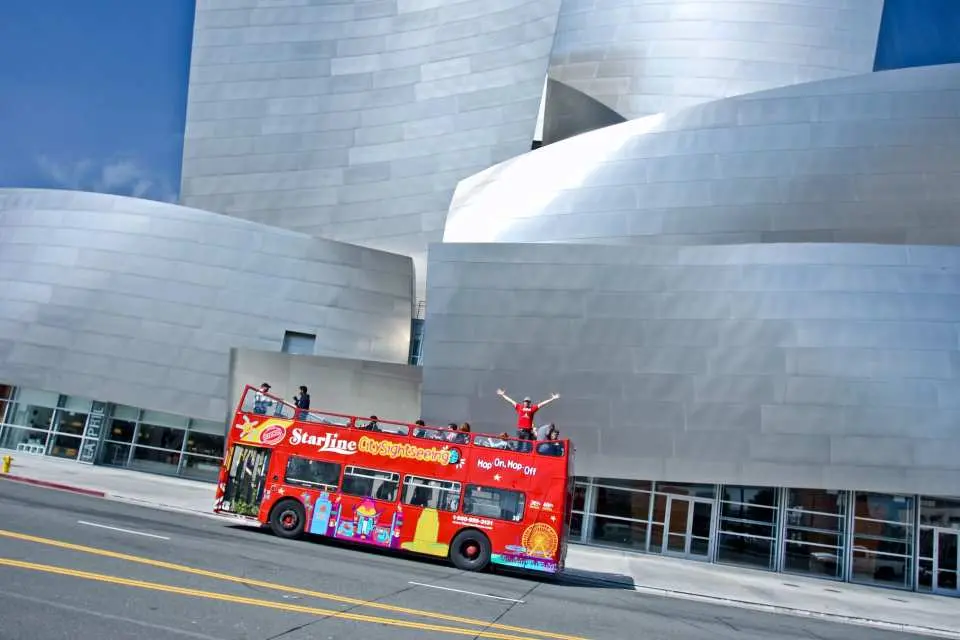 A red double-decker hop-on hop-off tour bus passing the modern Walt Disney Concert Hall in Los Angeles, offering flexible city exploration.