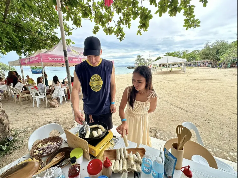 Participants and a local chef fry Filipino spring rolls during an outdoor beach cooking class in Siargao Island, enjoying an immersive culinary experience in a tropical island setting.