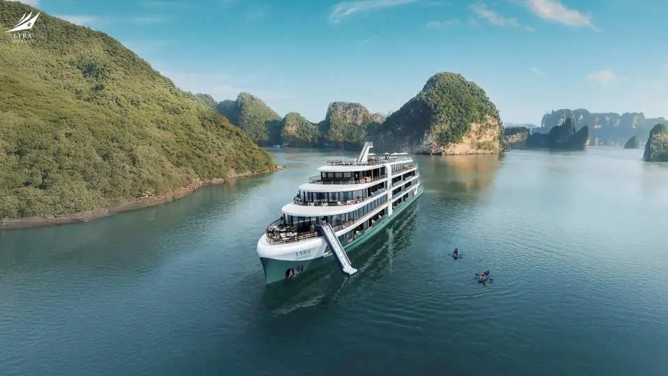An aerial view of the Lyra Grandeur luxury 6-star cruise gracefully sailing through Vietnam's Lan Ha Bay, surrounded by iconic limestone islands, with kayakers nearby.