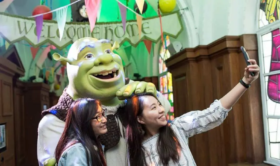 Two female tourists taking a selfie with a Shrek character at Shrek's Adventure! London, enjoying a fun, interactive family attraction.
