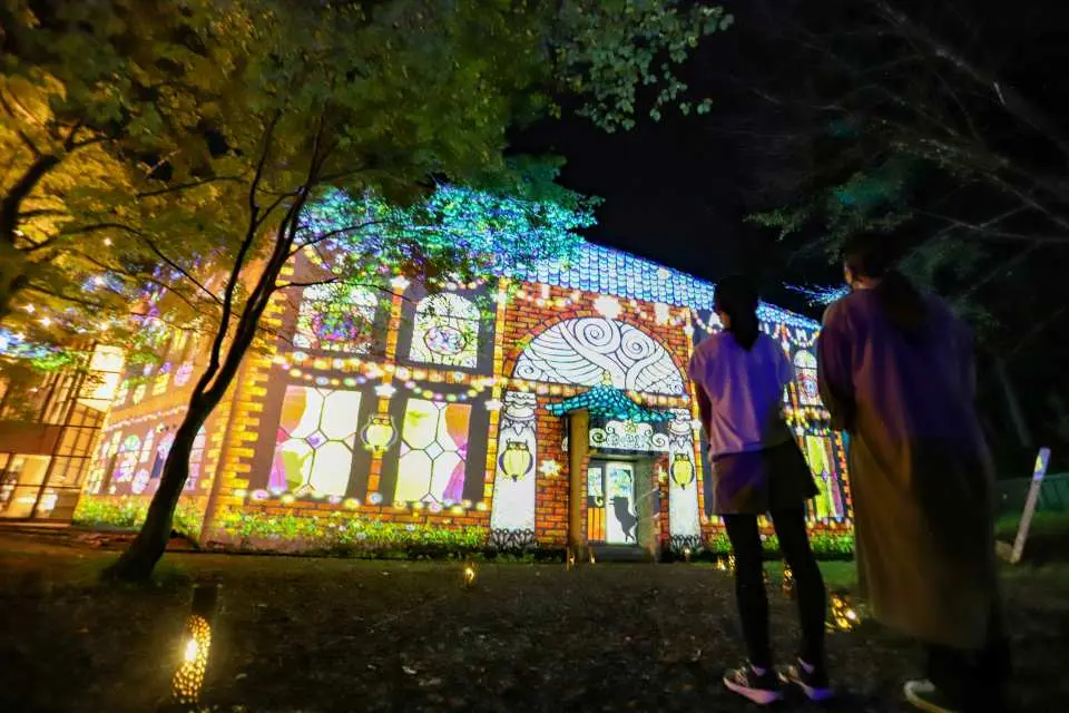 群马Chibam Land夜行中，两位游客背影望向被绚丽彩色光雕投影装饰的建筑，呈现彩绘玻璃般精致图案。