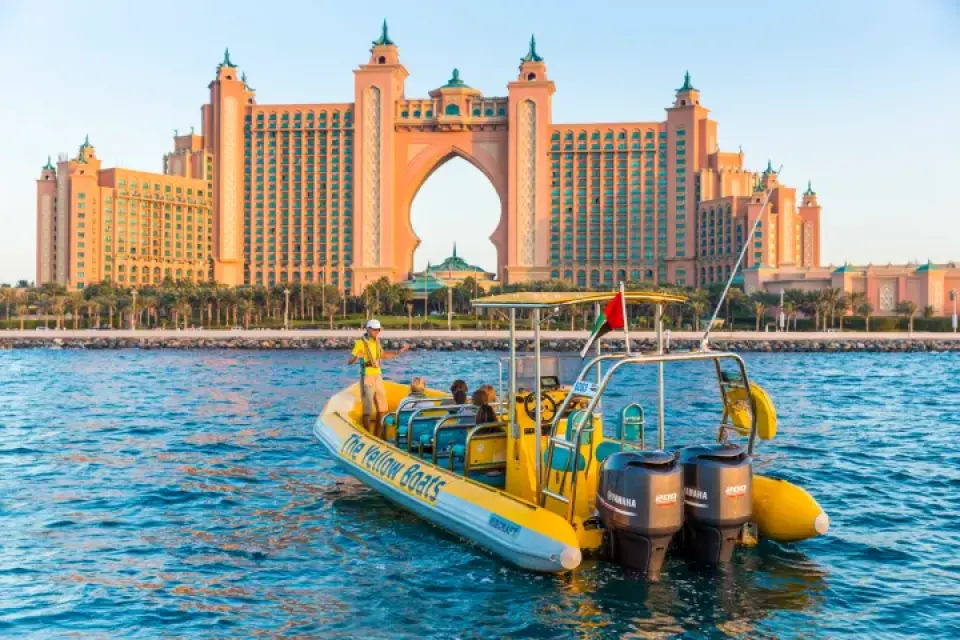 A yellow speedboat carrying tourists past the magnificent Atlantis, The Palm hotel, offering a unique perspective of Dubai's iconic landmark from the water on a sunny day.