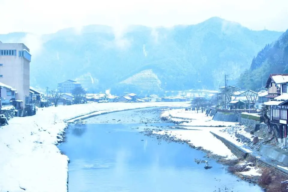 Panoramic winter view of Gujo Hachiman old town, featuring a clear river, traditional Japanese buildings, and distant snow-capped mountains under a bright sky.