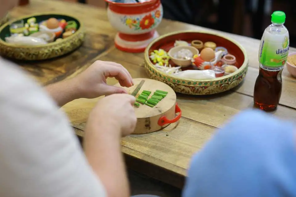 曼谷泰國菜烹飪課，學員們專注於切蔥花等食材，背景擺放著預先準備好的各式香料與配料，展現細緻烹飪過程。
