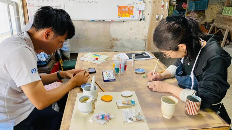 兩位個人，可能為指導老師與學員，在工坊共用工作台上專注地彩繪小型波佐見燒陶瓷磁鐵。