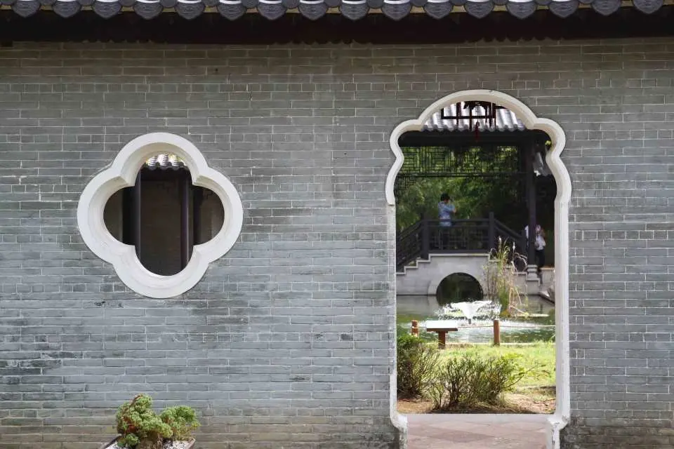 A decorative window and an arched gate in a traditional brick wall at Suwon Wolhwawon Garden lead visitors to admire the serene garden scenery, evoking a K-drama atmosphere.
