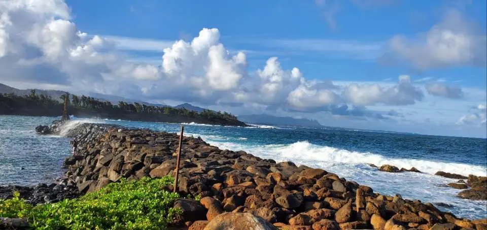 可愛島沿岸的崎嶇岩石防波堤，太平洋海浪拍打海岸，藍天白雲與綠色植物襯托出考艾島一日遊的自然野性之美。