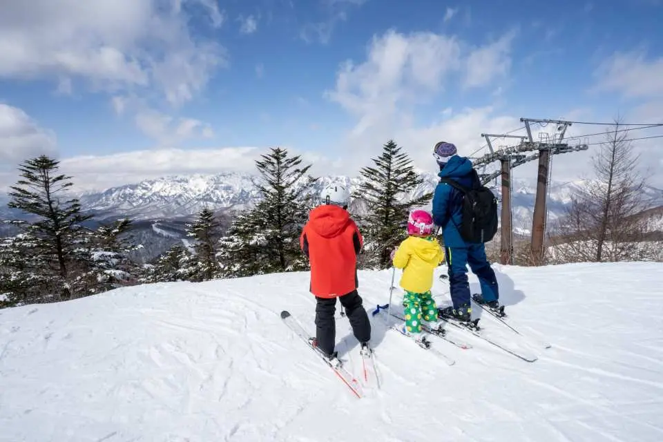 Family skiing at Togakushi Ski Resort. Parents and child standing at a snowy mountaintop enjoying panoramic winter mountain views under a blue sky.