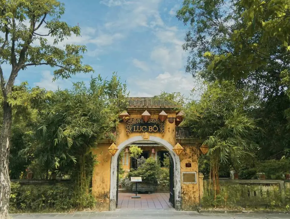 顺化传统工艺村内一座古老而优雅的拱形石门入口，门上装饰着传统灯笼，周围绿树环绕，充满历史气息与宁静。