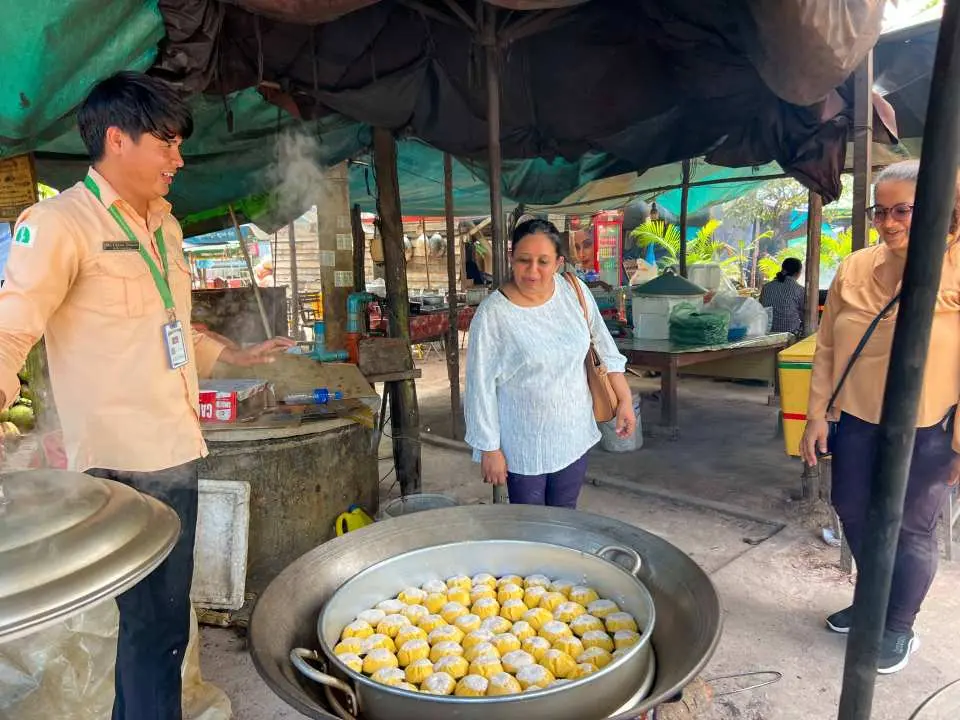 Local vendor preparing traditional palm cakes in a steamer pot at a palm cake village in Cambodia, with tourists observing the culinary process.