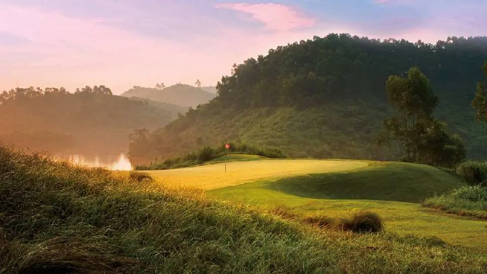 Lakeside green at Dongguan Mission Hills Golf Course, bathed in serene morning light or sunset glow, with water features and distant mountains, offering an elegant yet challenging tee-off experience.