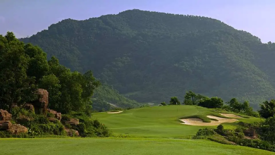 Distinctive fairway at Dongguan Mission Hills Golf Course, with a green and bunkers, flanked by unique rock formations and a majestic verdant mountain in the background, showcasing integrated design.