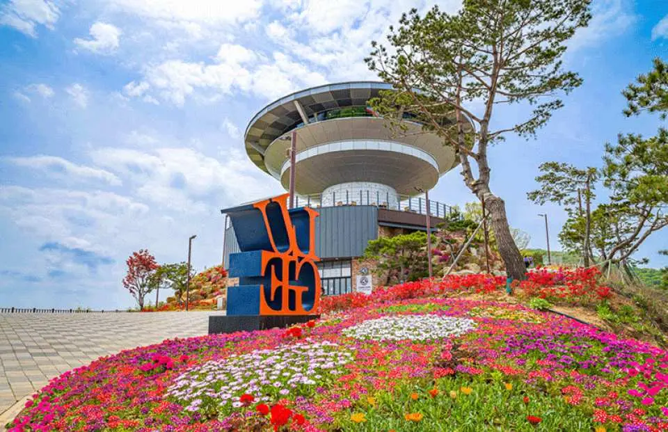 乌头山瞭望台现代建筑外观，周围环绕缤纷花卉与绿树，醒目的“ODUSAN”标志在蓝天下格外亮眼。