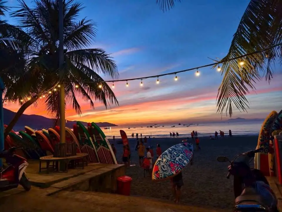 Stunning Da Nang Manta Been Beach at sunset or sunrise, showcasing colorful skies, palm trees with festive lights, and numerous stand-up paddleboards ready for the tranquil SUP adventure.