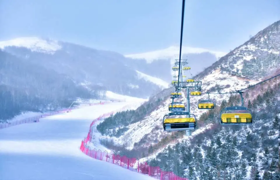 密苑雲頂樂園中，黃色纜車載著滑雪者，下方是寬闊平坦、設有紅色防護網的滑雪道，遠處群山環繞，適合初中級玩家。
