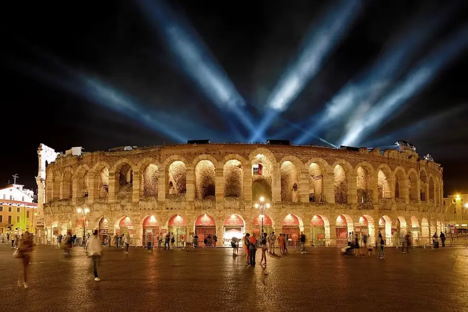 Captivating night view of the Verona Arena, illuminated by spotlights, showcasing ancient Roman ruins with visitors strolling in the piazza, offering free entry with the Verona Card.