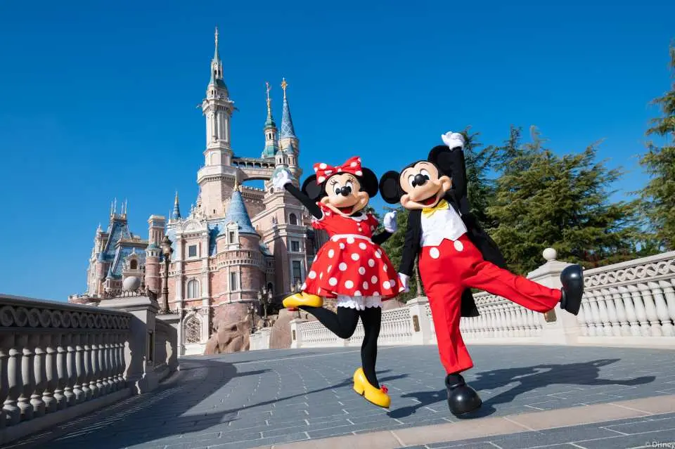 Mickey và Minnie mặc trang phục cổ điển, tạo dáng hoạt bát trước Lâu đài Câu chuyện cổ tích tại Disneyland Thượng Hải, với nền trời xanh trong.