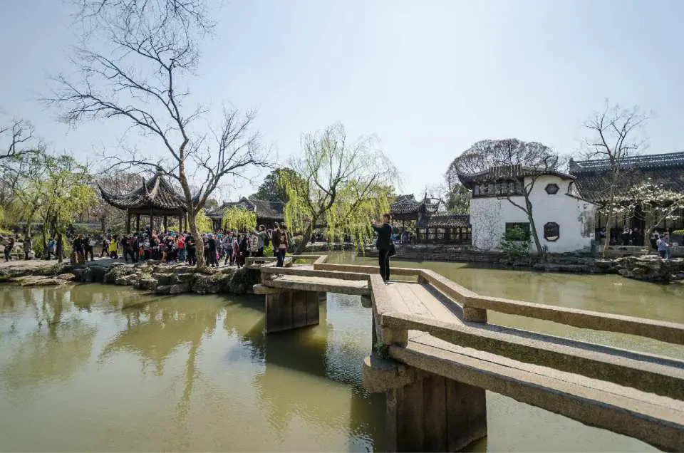 蘇州拙政園的廣闊湖景，遊客漫步於曲折木橋上，欣賞湖心亭與周遭古建築倒影，感受江南園林的開闊與精巧。