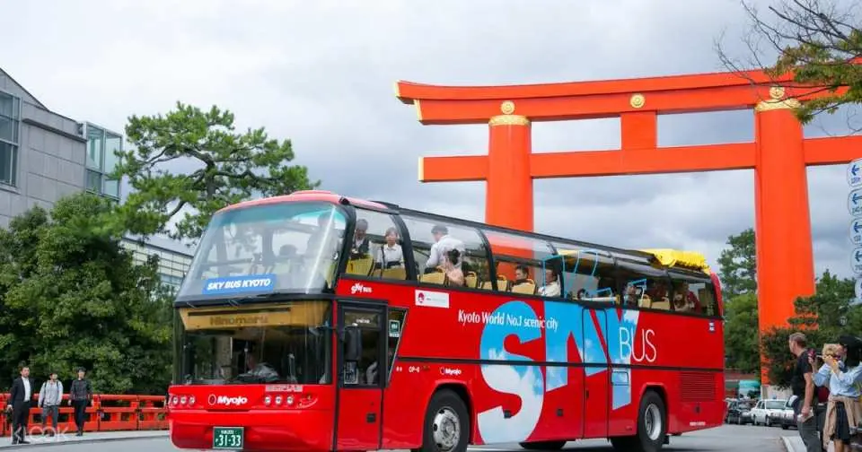 Kyoto's red double-decker open-top SKY BUS KYOTO passes a majestic red torii gate, with passengers enjoying panoramic city views and exploring cultural landmarks.