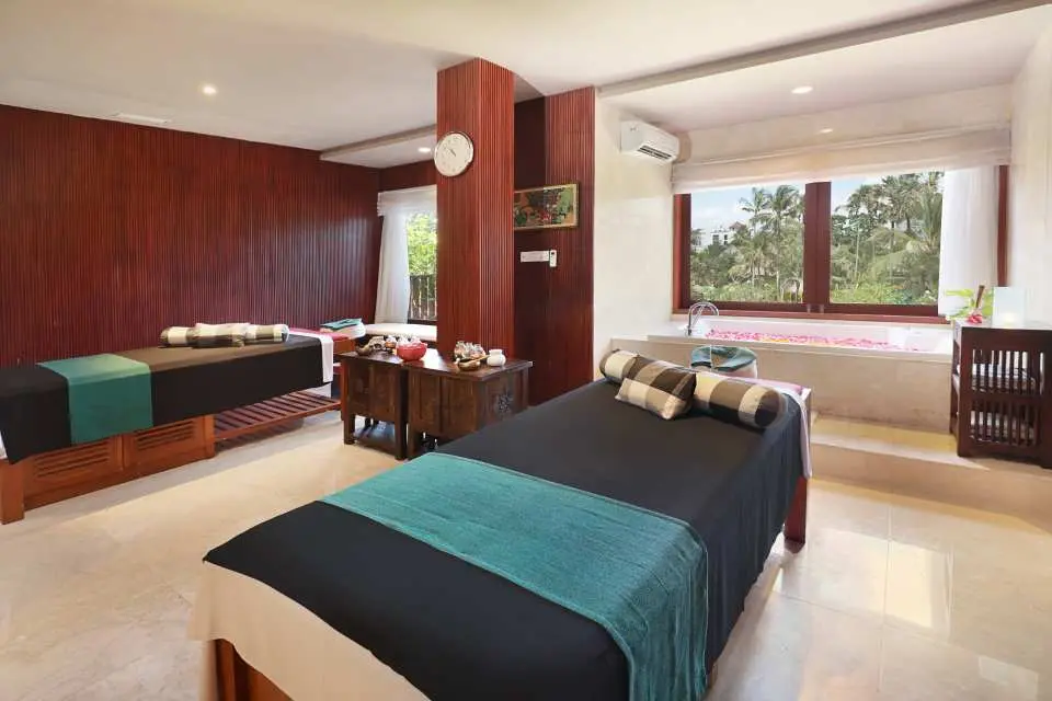 A serene couples' massage room at Usadha Spa in Bali, featuring two comfortable massage beds and a flower petal bath, offering a tranquil escape with tropical views outside the window.