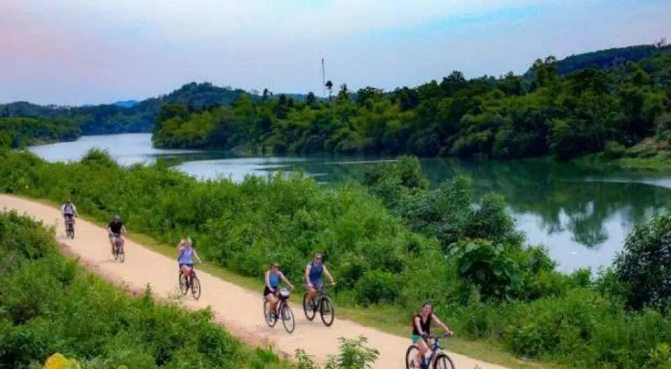 A group of tourists cycles along a scenic path by the Perfume River in rural Hue, enjoying lush mountains and river views.