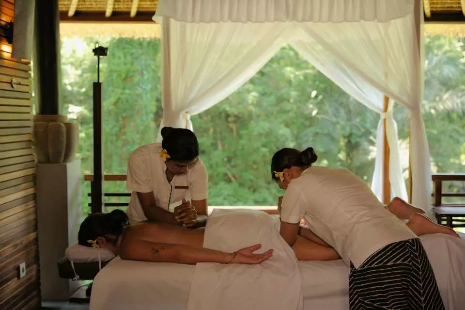 Two professional therapists performing a tandem massage on a guest at Maya Ubud Spa in an open-air treatment room, with Bali's lush rainforest visible outside.