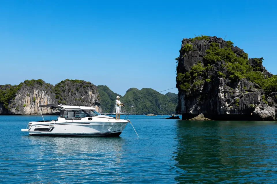 A man fishes from the bow of a private yacht in Halong Bay, enjoying a peaceful moment with dramatic limestone formations in the background, offering a serene escape.