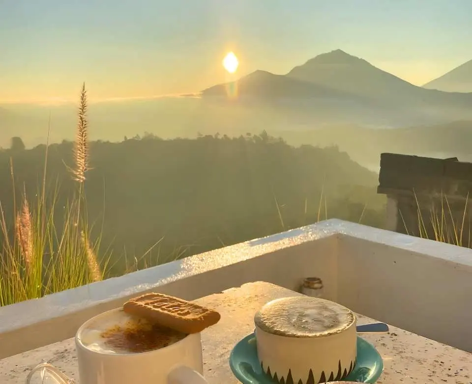 巴厘岛京那马尼咖啡馆，桌上两杯热饮与饼干，背景是巴图尔火山日出金黄色的天空和雾气缭绕的山峦。