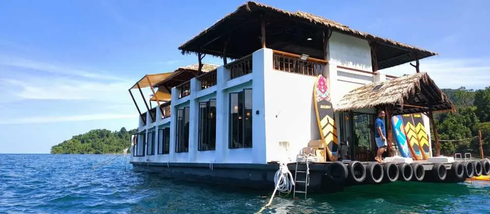 Side view of Gaya Island Floating Club in Kota Kinabalu, showcasing its thatched roof and wooden elements design, with a person relaxing on deck, surrounded by clear waters.