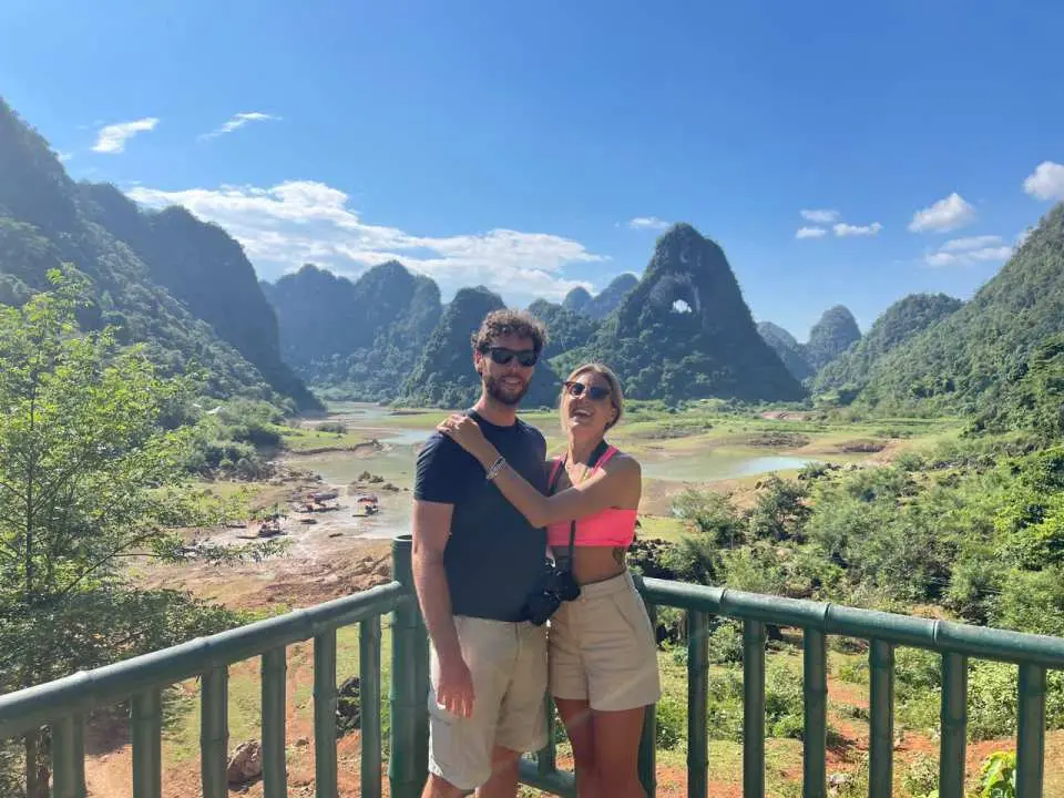 A couple posing by a wooden railing overlooking the emerald lake and distinctive karst mountains in the Angel Mountain (Núi Thủng) area, enjoying the natural scenery.