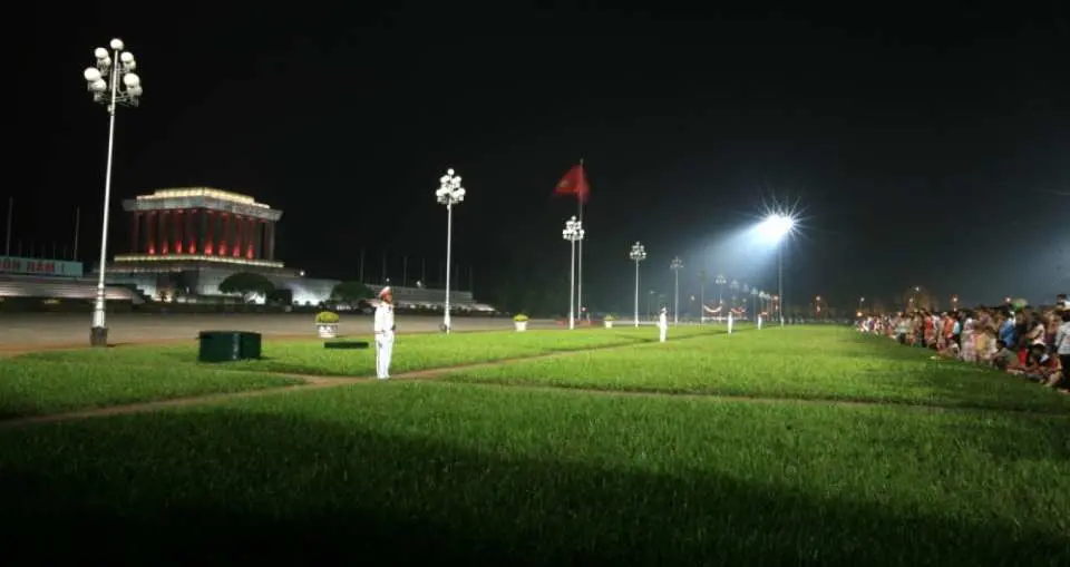 河内巴亭广场的广阔夜景，捕捉了灯火通明的胡志明陵寝和庄严的旗帜仪式，右侧有观众，共同见证这历史性的时刻。