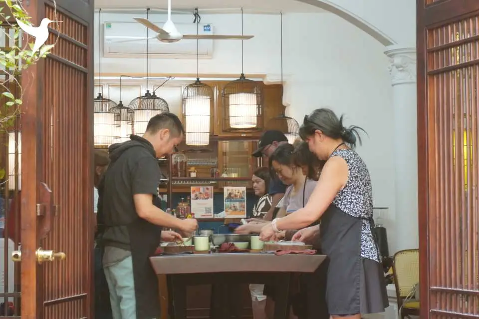 Participants are seen through a garden villa doorway, deeply engaged in a cooking class within Hanoi Rose Kitchen's bright and spacious indoor area, learning to prepare Vietnamese cuisine.