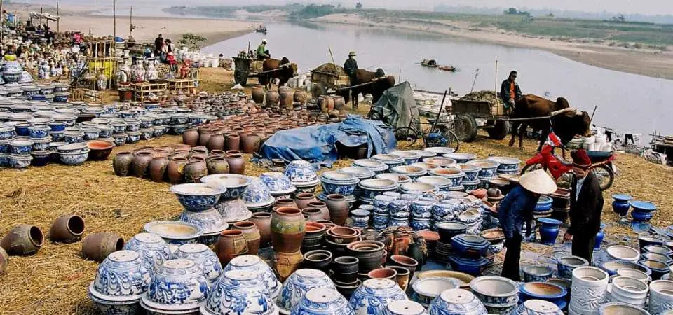 Chợ truyền thống sầm uất ven sông tại Làng gốm Bát Tràng, Việt Nam, với vô số sản phẩm gốm đẹp mắt được bày bán trên mặt đất và người dân địa phương giao thương bằng xe ngựa hoặc xe bò, phản ánh nhịp sống đời thường sống động.
