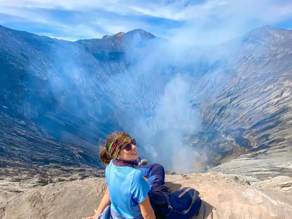 一位女性遊客戴著頭巾和墨鏡，坐在布羅莫火山火山口邊緣，遠眺冒煙的火山深淵。
